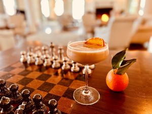 A stylish cocktail garnished with an orange slice sits beside a chessboard in a cozy living room.